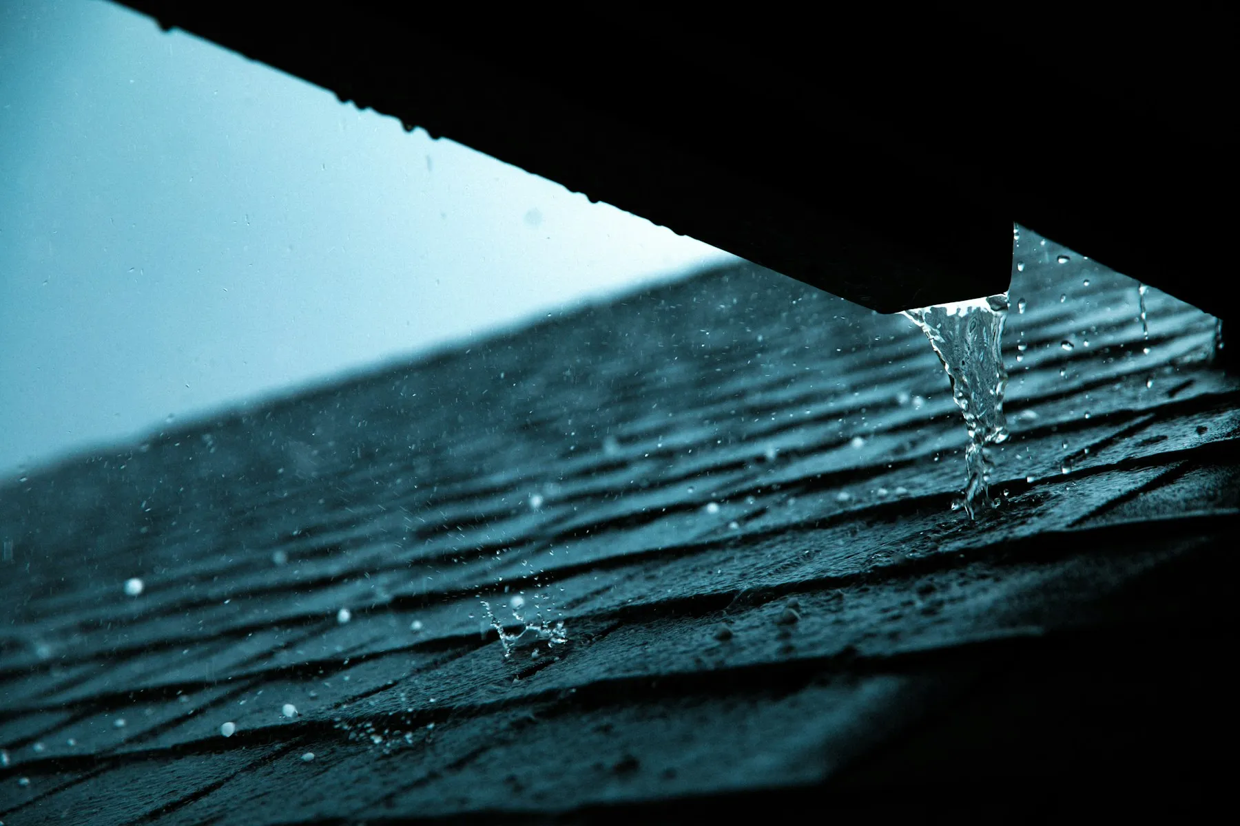 Rainwater pouring from the end of an eavestrough onto dark asphalt shingles during a storm.