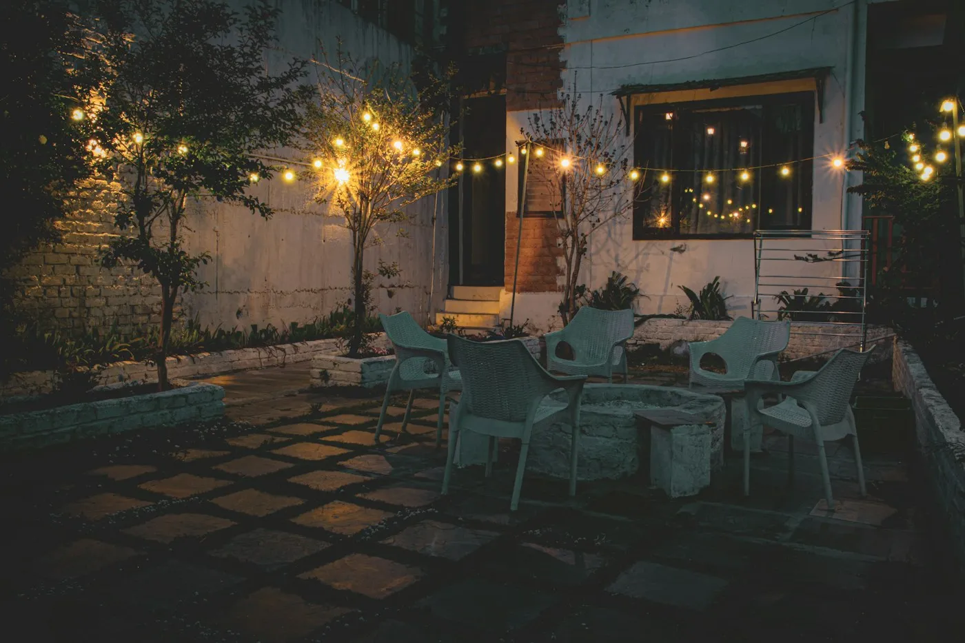Bistro bulbs strung over a back patio at night, lighting the table and the small trees around it.