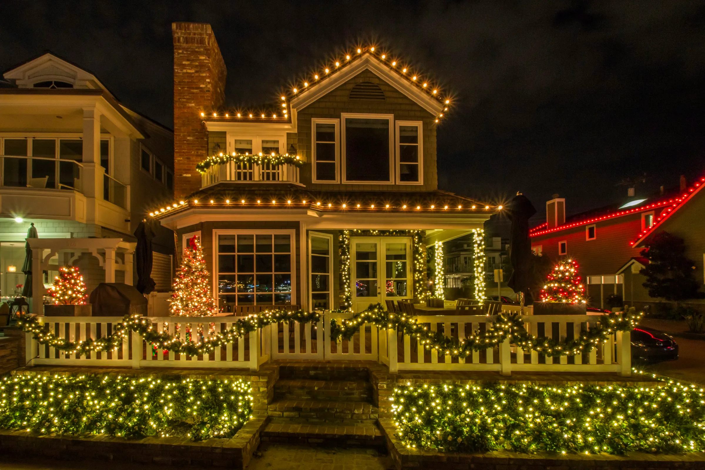 A two-storey home at night with warm white bulbs tracing every roof edge, dormer and porch line, with lit garland along the front fence.